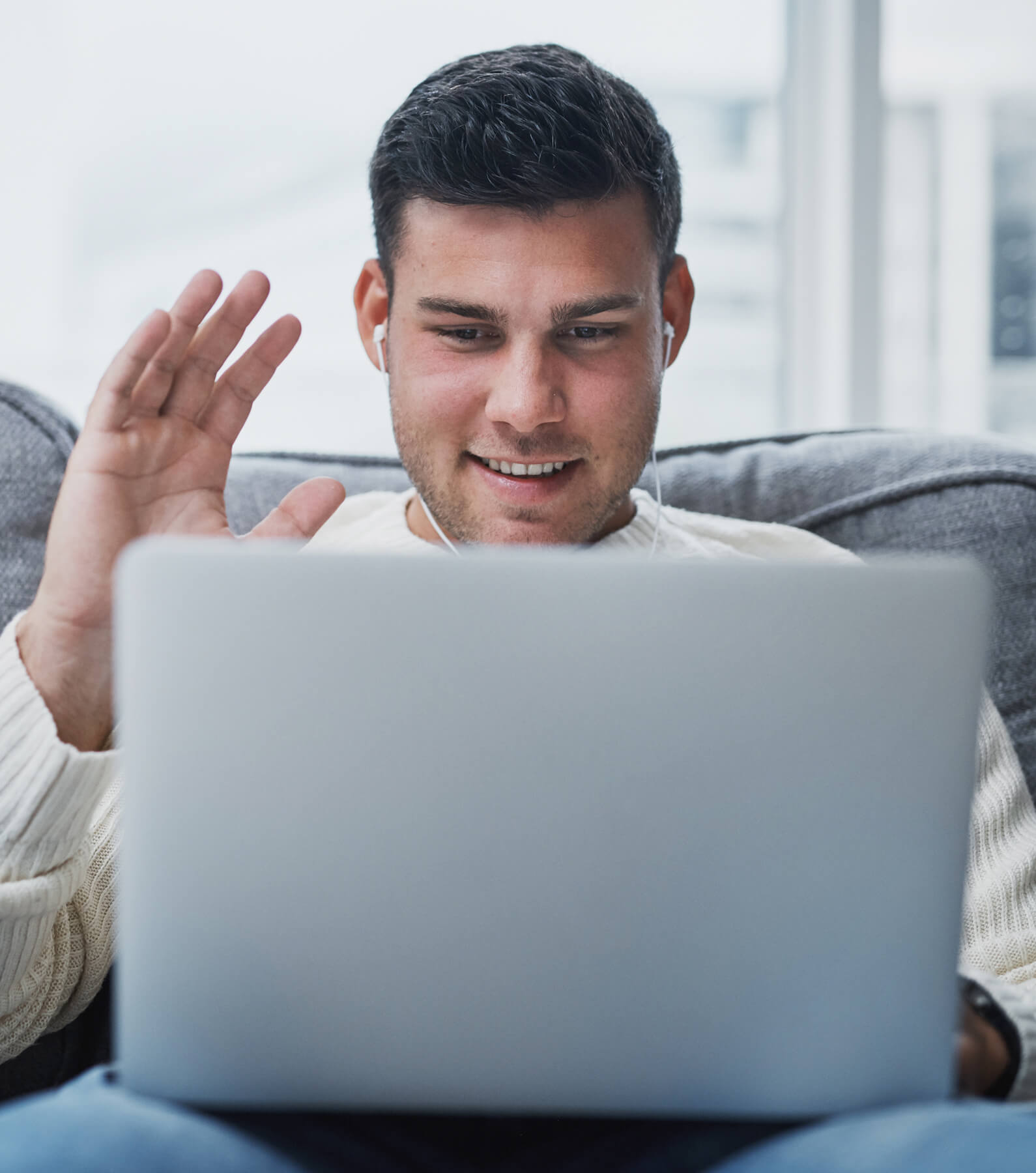 A man wearing earphones sits on a couch, looking at a laptop and waving—perhaps connecting with colleagues to discuss physician burnout during a video call.