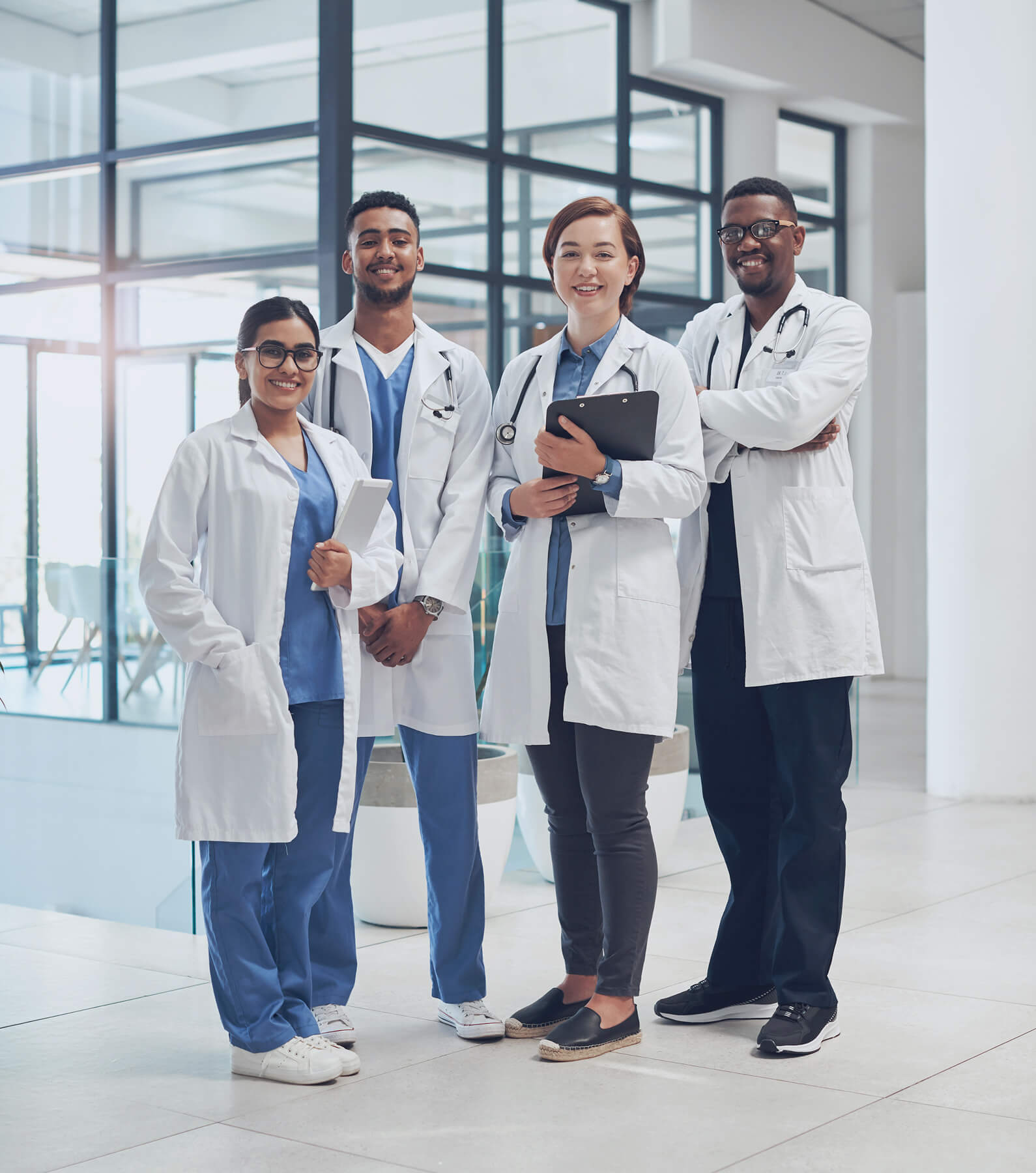 Four doctors in lab coats stand together in a modern medical facility, smiling and looking at the camera. Their teamwork reflects a supportive environment that helps prevent physician burnout. One holds a clipboard.