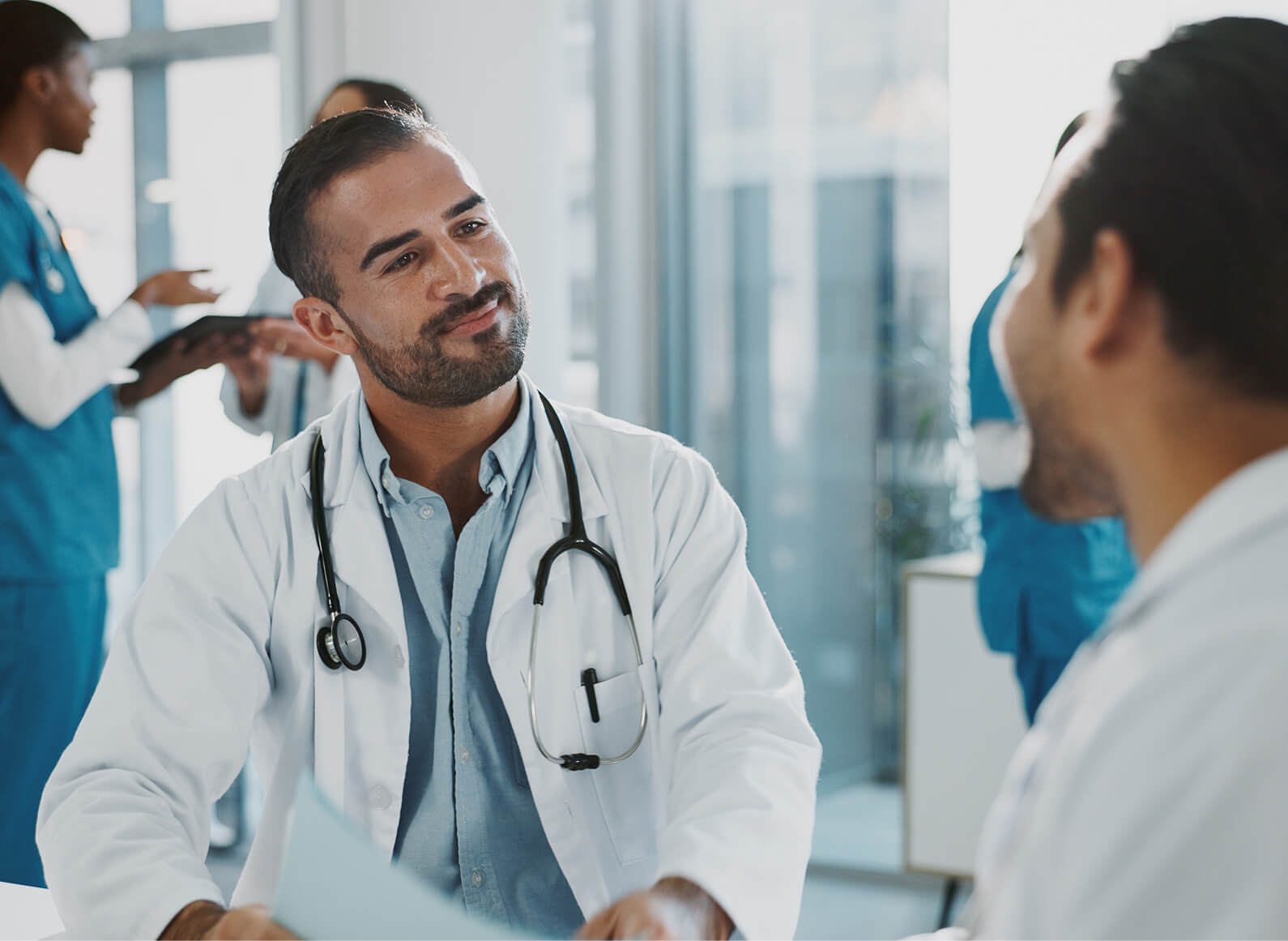 A doctor with a stethoscope around his neck discusses physician burnout with a colleague in a bright medical office, while two other healthcare professionals work in the background.