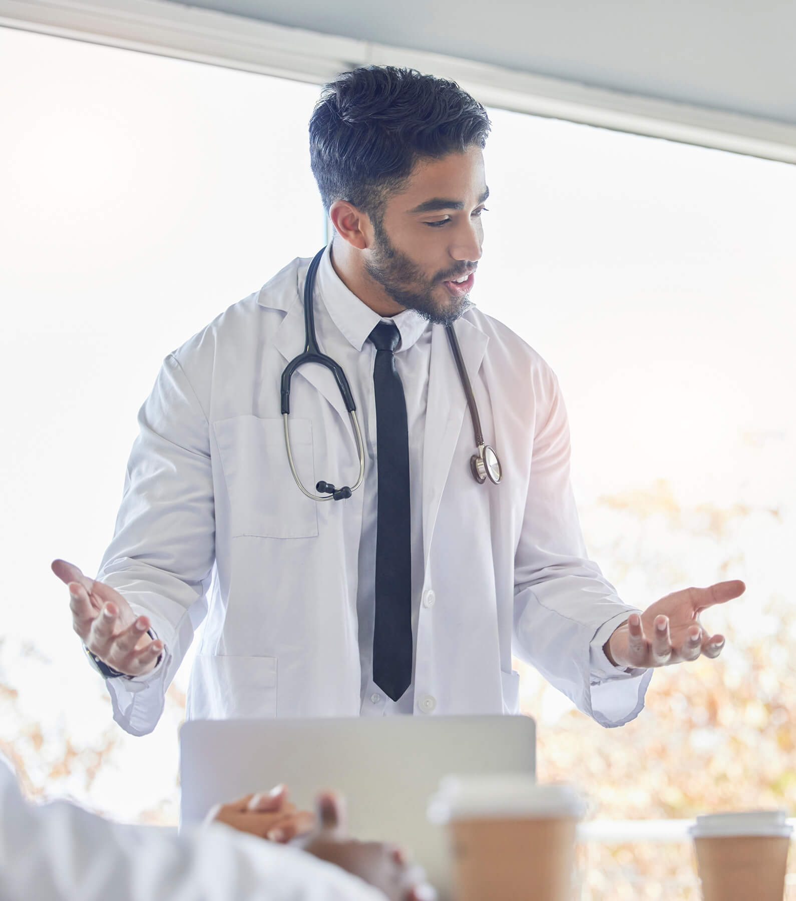 A male doctor, wearing a white coat and stethoscope, gestures with his hands while speaking about physician burnout in a bright, indoor setting.