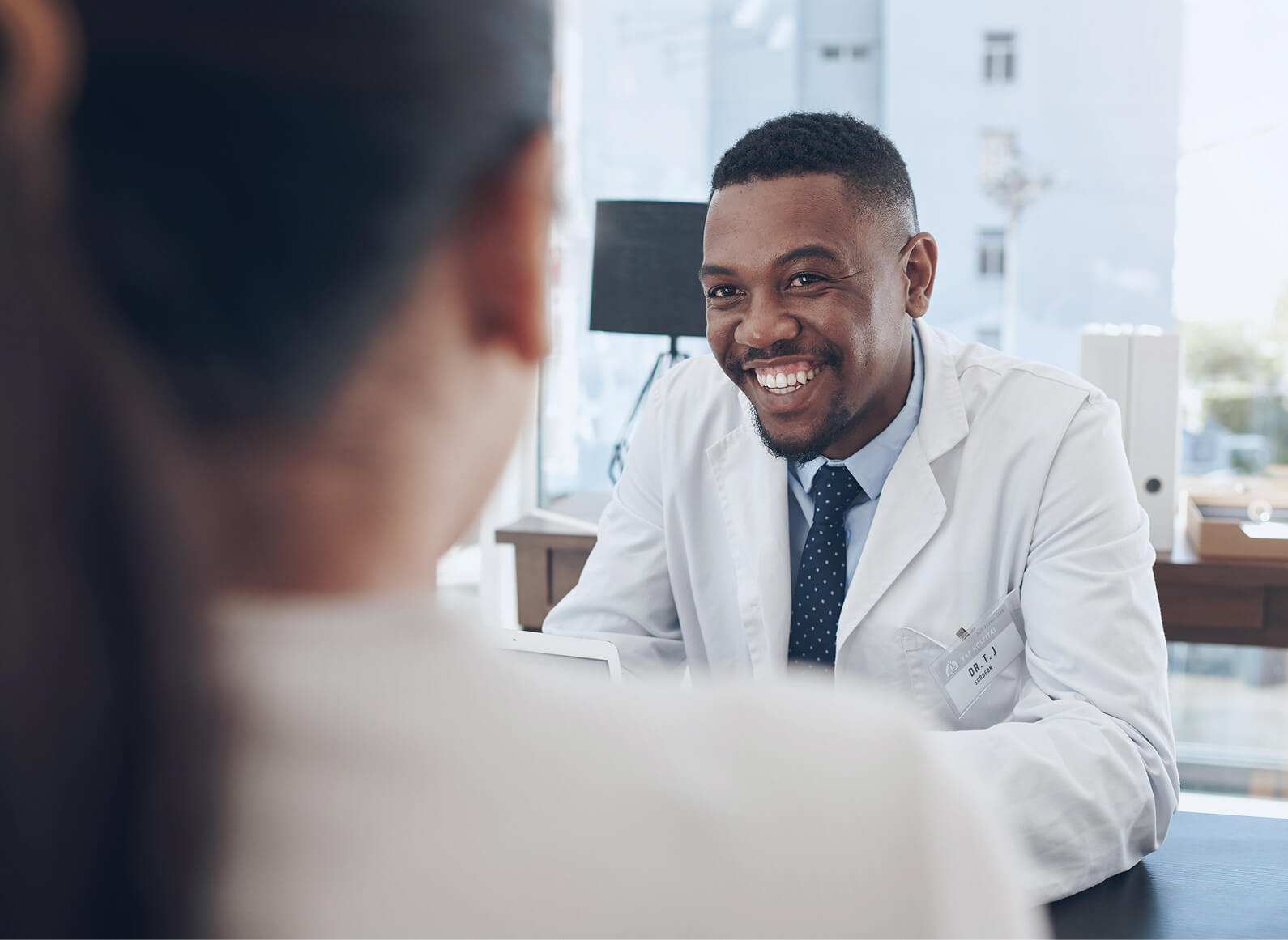 A doctor in a white coat smiles while talking to a patient across a desk in a brightly lit office, fostering connection and helping prevent physician burnout.