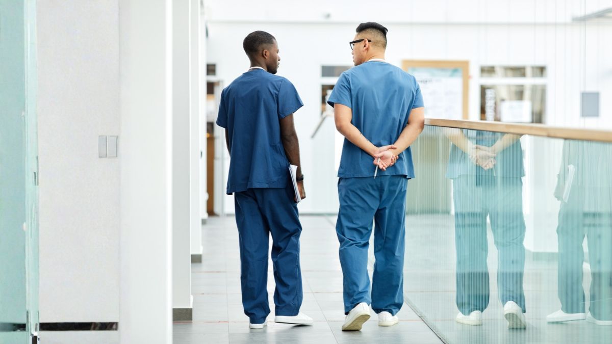A senior physician walks alongside a younger physician. The senior doctor handles the curbside on his terms and as if pressure is a privilege.